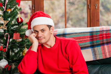 Young man sitting on the couch with santa hat in deep thoughts at home, looking sad at camera, over christmas tree background on living room.の写真素材