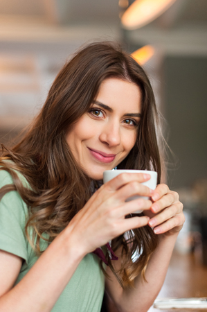 Beautiful girl taking a break sitting at bar in the cafe shop, smiling happy holding a cup of coffee and looking at camera.の写真素材