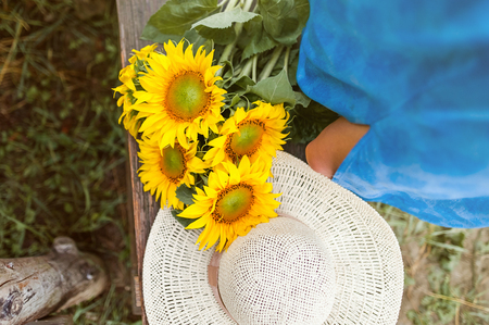 Top view of a sun flower bouquet and a straw hat on a wood banch and a woman standing next to it, wearing a blue dress.の写真素材