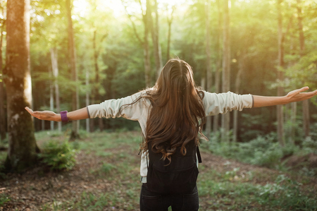 Young woman emgracing the sun and nature with backpack in the forest on sunset light in the autumn season, looking up, with her hands in the air, exploring the nature.の写真素材