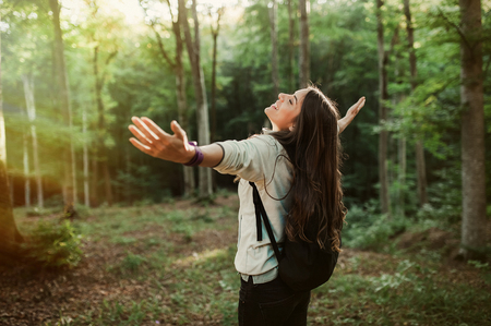 Young woman emgracing the sun and nature with backpack in the forest on sunset light in the autumn season, looking up, with her hands in the air, exploring the nature.の写真素材