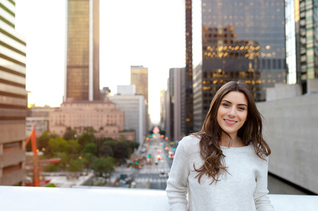 Beautiful young woman sitting on a bridge across the boulevard in urban scenery, downtown, at sunset, smiling at camera.の写真素材
