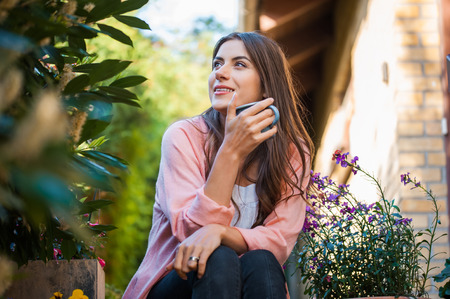 Relaxed young girl smiling and looking side, sitting on home terrace, holding a mug drinking a hot beverage Hobby and resting leisure concept.の写真素材