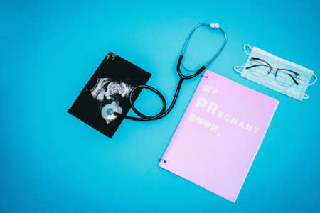 Stethoscope, glasses, medical mask, ultrasound picture of a child and pregnant book on a light blue background. Flat-lay, top view with place for your text. Medical trends and concept.の写真素材