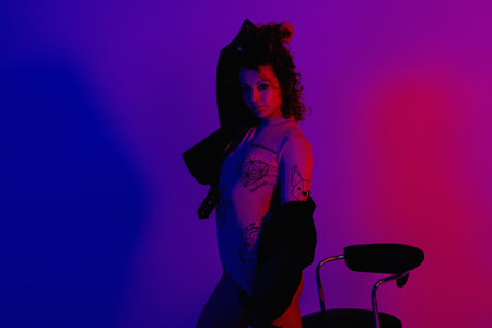 Fashion model in stylish clothes at the neon lights in the studio. Studio portrait of a sexy woman in a leather jacket, bodysuit and boots on a red, blue, purple ultraviolet background in a nightclubの写真素材