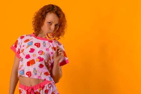 Studio photo of a beautiful red-haired woman in pink pajamas on a yellow background. The woman is sucking a lollipop and looking at the camera. Place for your text.の写真素材