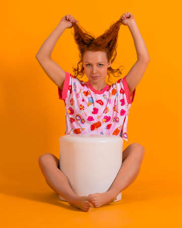 Studio photo of a beautiful red-haired woman in pink pajamas on a yellow background. A woman sits by the armchair clutching her hair and looks at the camera.の写真素材