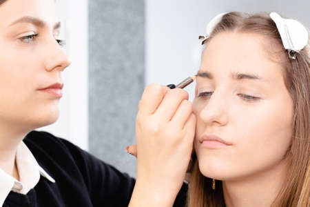 Professional makeup artist applies makeup to a beautiful girl model. The face of girl with clean skin. Real life Makeup and Cosmetology concept. Makeup artist draws eyebrows with a pencil to the girlの写真素材