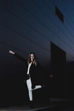 Sexy girl posing against a dark wall in the evening. An elegant fashionable girl in a long black coat, white jeans, with a leather bag stands near the wall of the building. Beautiful girl for a walkの写真素材
