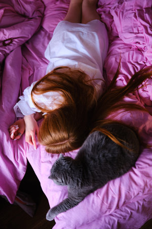 pretty young ginger woman with her gray british cat in pink bed at home girl in white shirt shot...