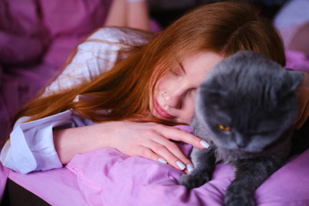 pretty young ginger woman with her gray british cat in pink bed at home girl in white shirt hug the catの写真素材