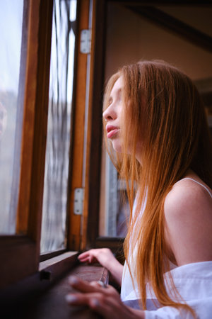 Young redhead Woman in white shirt Looks Out The Window on the balcony Deep In Thought red hairs...
