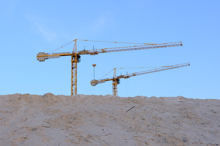 Two construction cranes tower over a mound of sand against a backdrop of blue sky. Perfect for illustrating construction progress and industrial developmentの写真素材