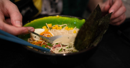 Man enjoys a delicious bowl of ramen, featuring corn, seaweed, noodles, eggs, and meat. Close-up...