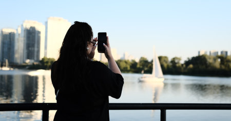 young woman in black films stunning river scene on her phone, featuring sailboat white sails modern residential buildings in the background. Perfect showcasing urban beauty peaceful waterfront views.の写真素材