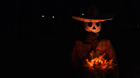 A Halloween character with a pumpkin body and a white skull-like face wearing a hat, red shirt, and sweater holds glowing autumn yellow leaves in hands, creating an eerie and festive atmosphere.の写真素材
