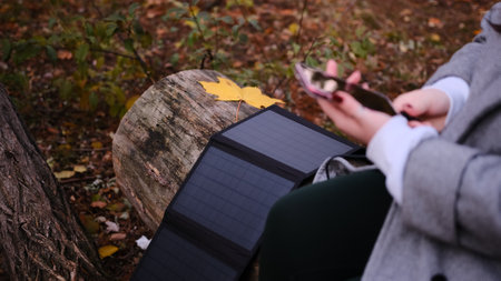 A woman in a gray coat charges her phone using a portable solar panel in a serene forest setting. The scene highlights sustainable technology, off-grid living, and eco-friendly solutions in nature.の写真素材