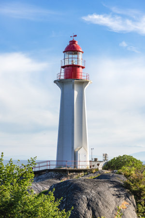 Red and white lighthouse surrounded by green bushes with ocean and mountains on the backgroundの写真素材