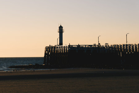 Old lighthouse on the pier with people admiring sunset