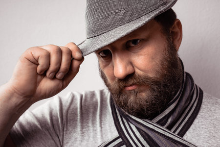 Bearded young hipster man wearing gray hat and scarf, fashion portrait against gray wall.