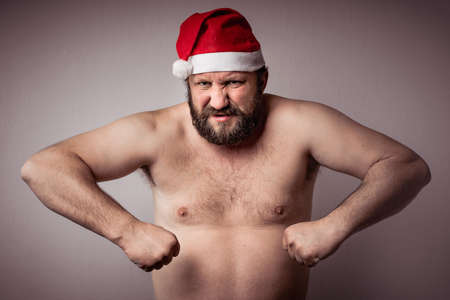 An Angry bearded half-naked Santa Claus man with a long beard in Christmas or Xmas new year hat looking straight into camera, anger, rage.の写真素材