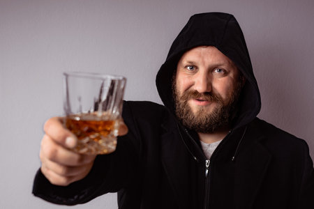 Handsome elegant bearded man wearing black coat with hood drink and toast with some strong liquor at...