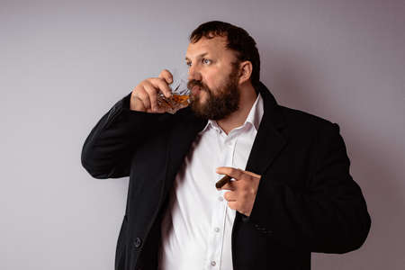 Handsome confident mature man with beard who wearing coat and shirt drink some strong liquor and smoking his cigar at celebration.の写真素材