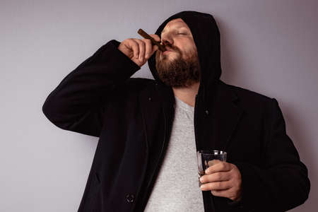 Handsome elegant bearded man wearing black coat with hood drink some strong liquor and smoking his cigar at celebration.の写真素材