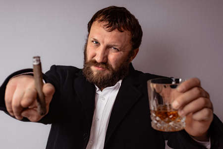 Bearded man in modern coat and shirt smoking his cigar and drink some strong liquor pointing with finger.の写真素材