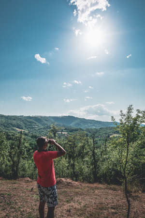 A man uses his binocular and looking over the trees and mountain hill.  A man in the red t-shirt looks at a beautiful landscape with binoculars.の写真素材