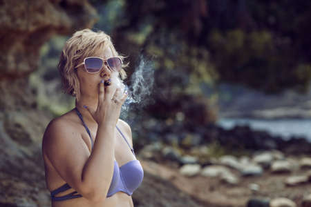 Blonde woman smoking a cigar. Blonde woman in bikini smoking a cigar at the beach.の写真素材