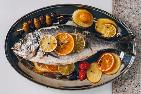 Sea gilt-head bream fish on the plate baked with potatoes, rosemary, lemon, orange, olives, tomatoes and lime. Fresh Orata, Dorade fish preparation.の写真素材