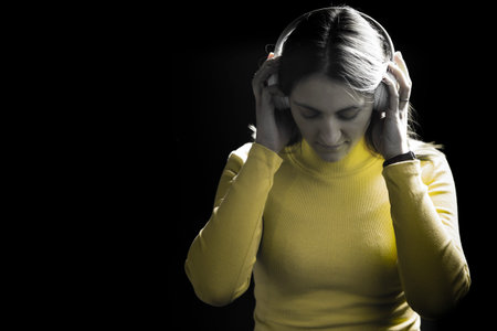 Portrait of a young woman with dark hair in a yellow t-shirt with headphones on her ears on a black...