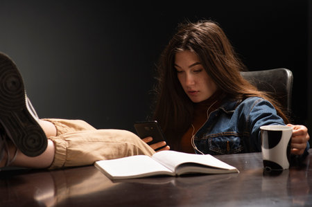 Female student sitting with her feet on a desk and a message key on a mobile phoneの写真素材