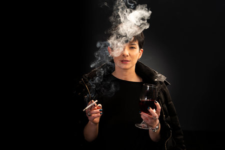 A young woman drinks wine and smokes a cigarette on a black backgroundの写真素材