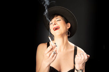Young woman with a cigarette in a black hat on a black background. Concept of bad emotions.