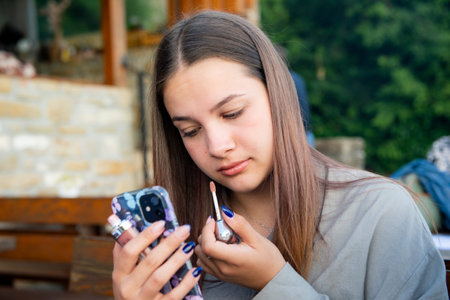 Young girl applying lipstick while looking at herself in the mirror of a mobile phone outdoorsの写真素材