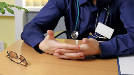 A medical consultant sits at a table and listens to someone. His hands are clasped and he...