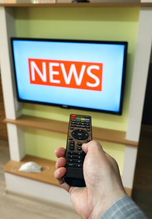 Hand holds the remote control and directs it to the TV, changing channels. News on the TV screenの写真素材