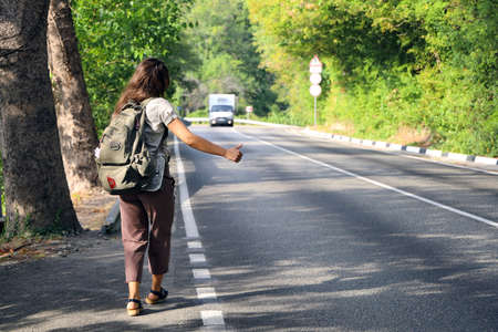 Young hitchhiker with backpack behind her back stops an approaching van with her thumb raised on sunny summer day outside the cityの写真素材