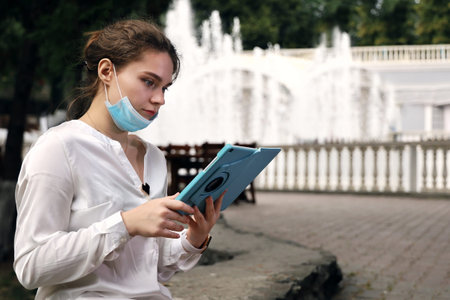 Young woman student in medical facial mask sits in quiet, deserted place in the park against the...