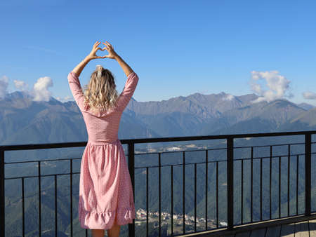 Young woman in pink dress and blond, curly hair stands with her back near the railing of the observation deck, looks at the mountains and makes heart sign with her hands, on summer sunny dayの写真素材