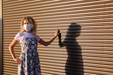 Young woman in medical face mask stands near the closed street shutters. She touches her shadow with her hand and looks away. Concept of loneliness during pandemicの写真素材
