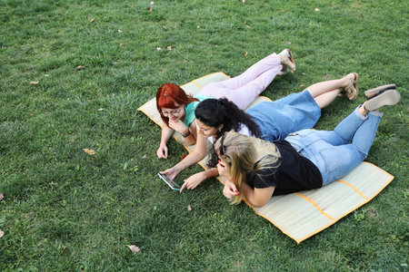 Three young women friends lie on the blanket on the grass and look at the smartphone screen. They...