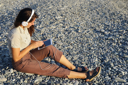 Young woman in white headphones holds tablet in her hands and chooses something while sitting alone...