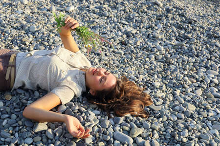 Young smiling woman laying on pebble and holds small bouquet of wildflowers. Concept of serenity and happiness, with copy spaceの写真素材