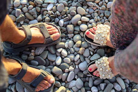 Male and female feet of lovers in sandals opposite each other on pebble surface, top view
