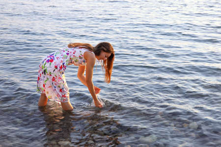 Young smiling woman in summer dress stands barefoot in water and washes her hands. Outdoors at sunset with copy spaceの写真素材