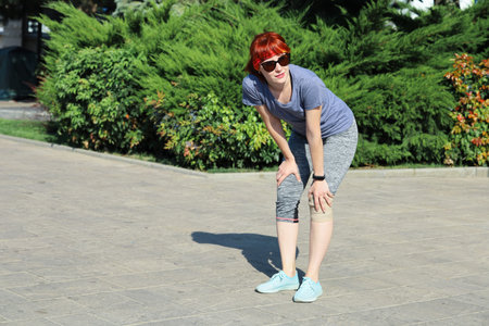 Young red-haired smiling woman in sportswear stands outdoors, resting during sports, supporting...