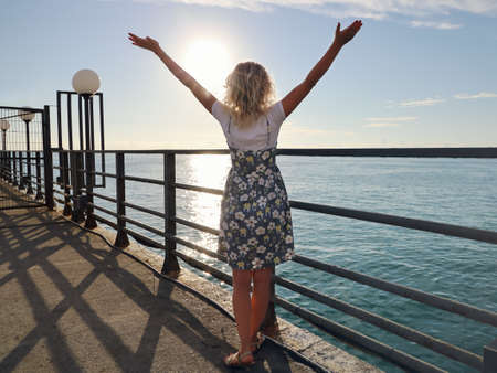 Young woman in summer dress stands back with raised hands, on deserted pier on background of sea and sky in sunsetの写真素材